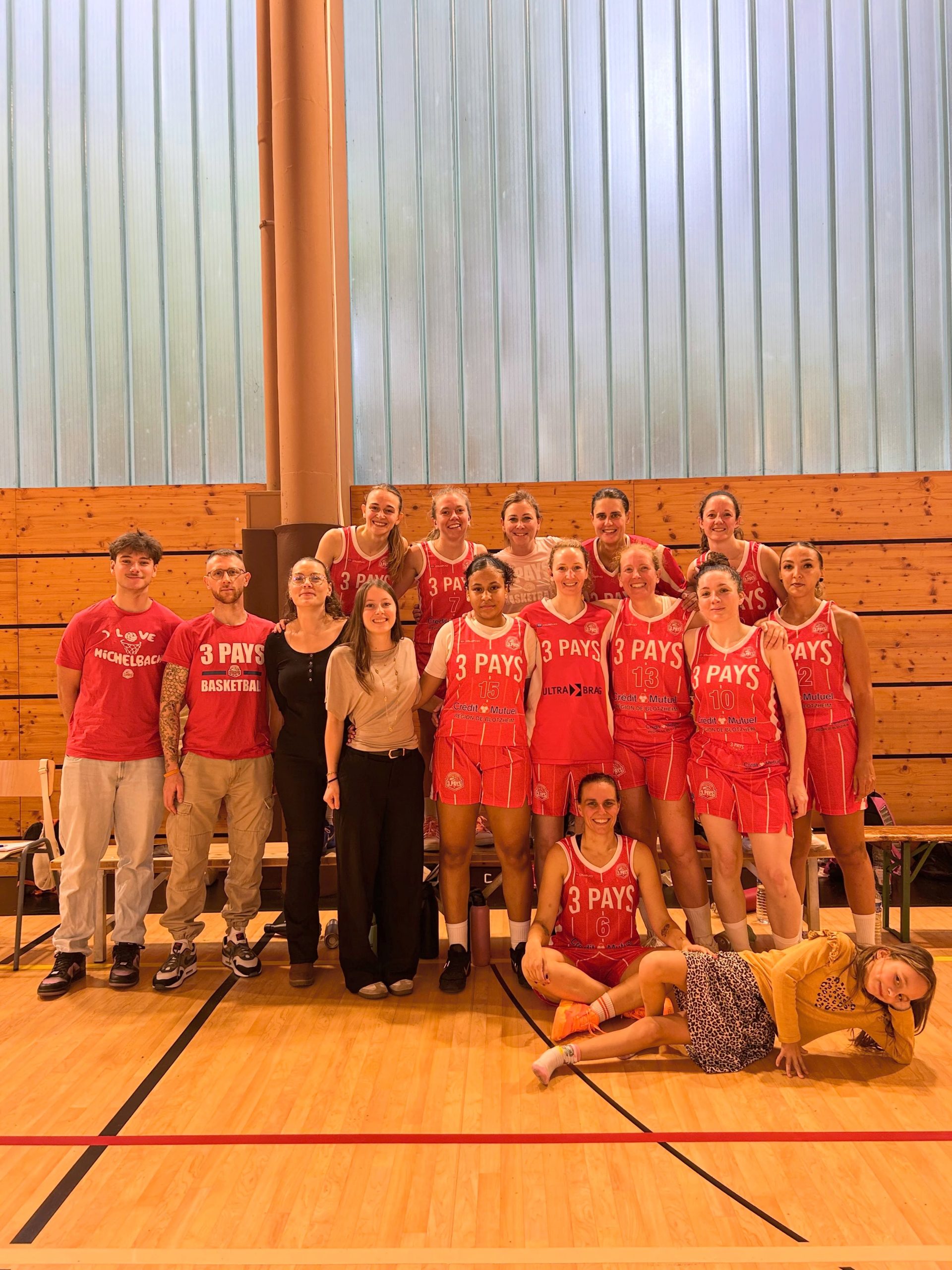 Joueuses adultes en maillots rouges, réunies avec leur encadrement dans une salle de basket.